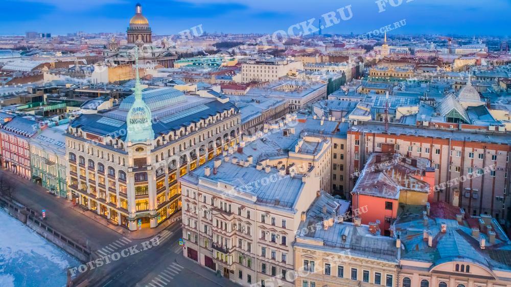 Это фотография, на которой запечатлен день или ранний зимний вечер в центре Санкт-Петербурга. Изображение сделано с высоты, что позволяет увидеть плотную застройку исторической части города с множеством зданий, выполненных в классическом стиле. В центре внимания — хорошо освещенное здание с куполом и зеленой крышей, выделяющееся на фоне окружающих его более тусклых фасадов. Этот снимок может быть использован для иллюстрирования путеводителей, исторических и культурных обзоров города или как рекламный материал для туристических агентств.
